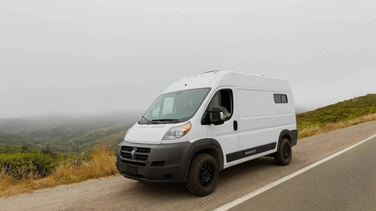 White Ram ProMaster high-roof camper van conversion by The Vansmith, parked roadside at a foggy hillside overlook, black off-road tires and fender flares visible