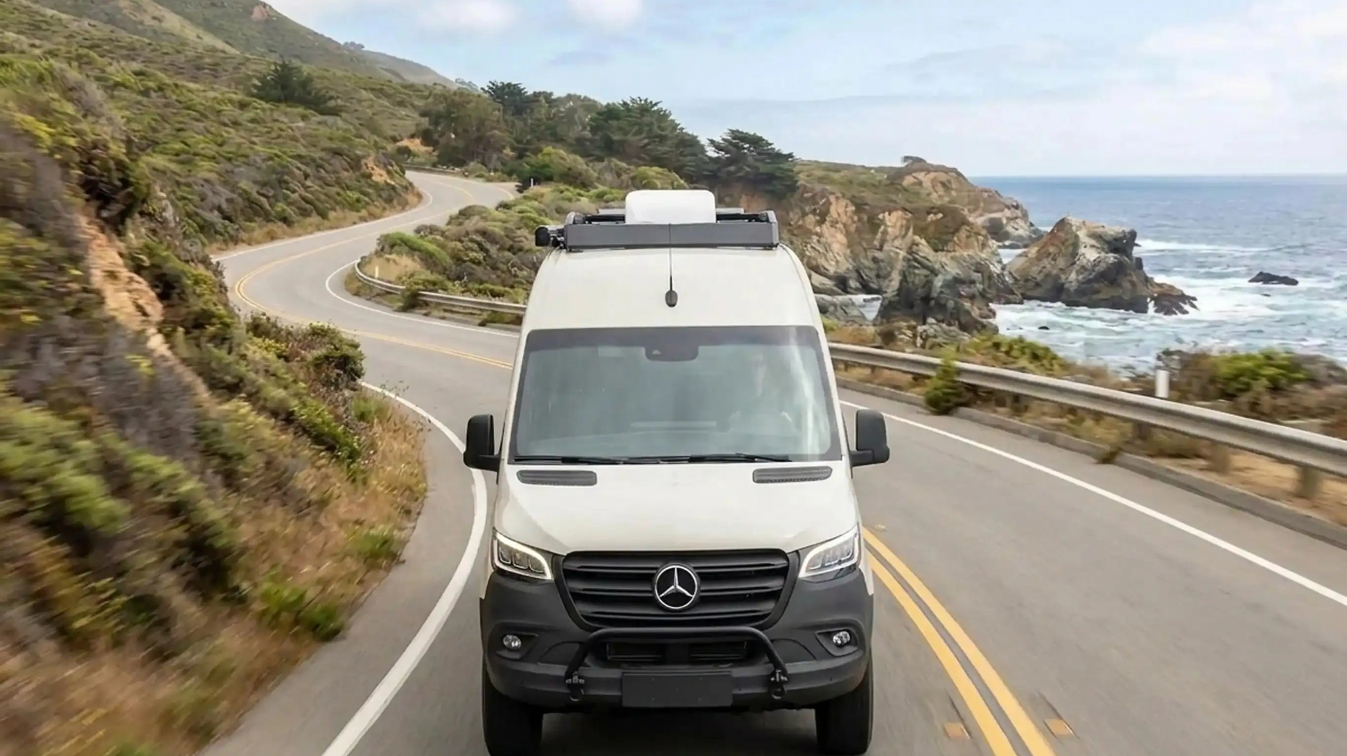 White camper van driving on winding coastal road with ocean and cliffs in the background