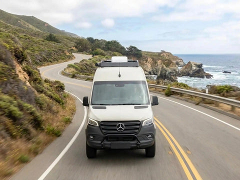 White camper van driving on winding coastal road with ocean and cliffs in the background