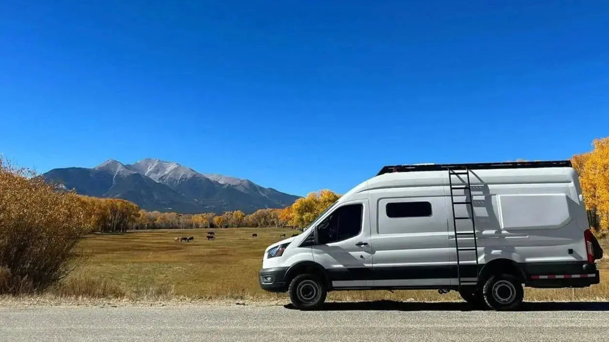 White Ford Transit camper van conversion with roof rack and ladder, parked roadside against a Colorado mountain backdrop