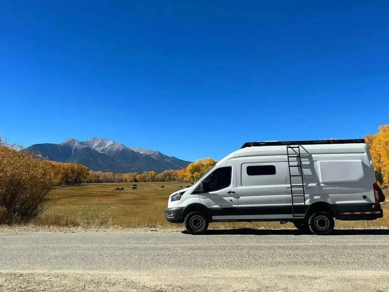 White Ford Transit camper van conversion with roof rack and ladder, parked roadside against a Colorado mountain backdrop