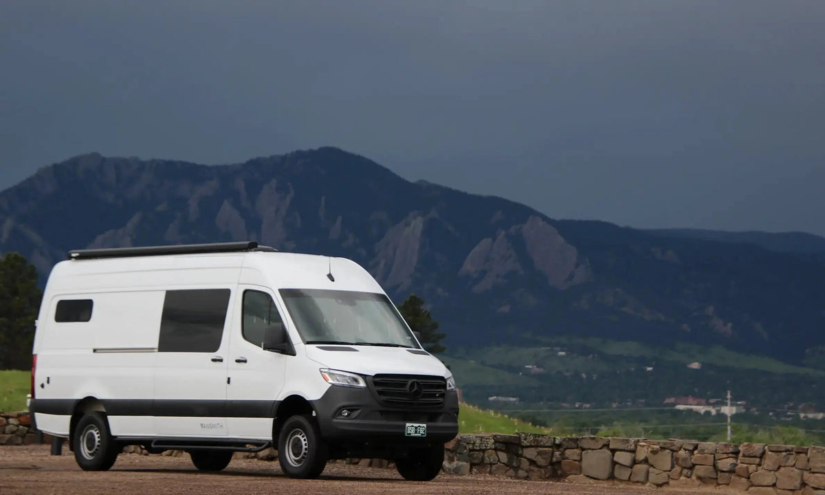 White Mercedes-Benz Sprinter 4x4 camper van built by The Vansmith, parked on a gravel pull-off with Boulder, Colorado's Flatirons and a storm-lit sky behind it