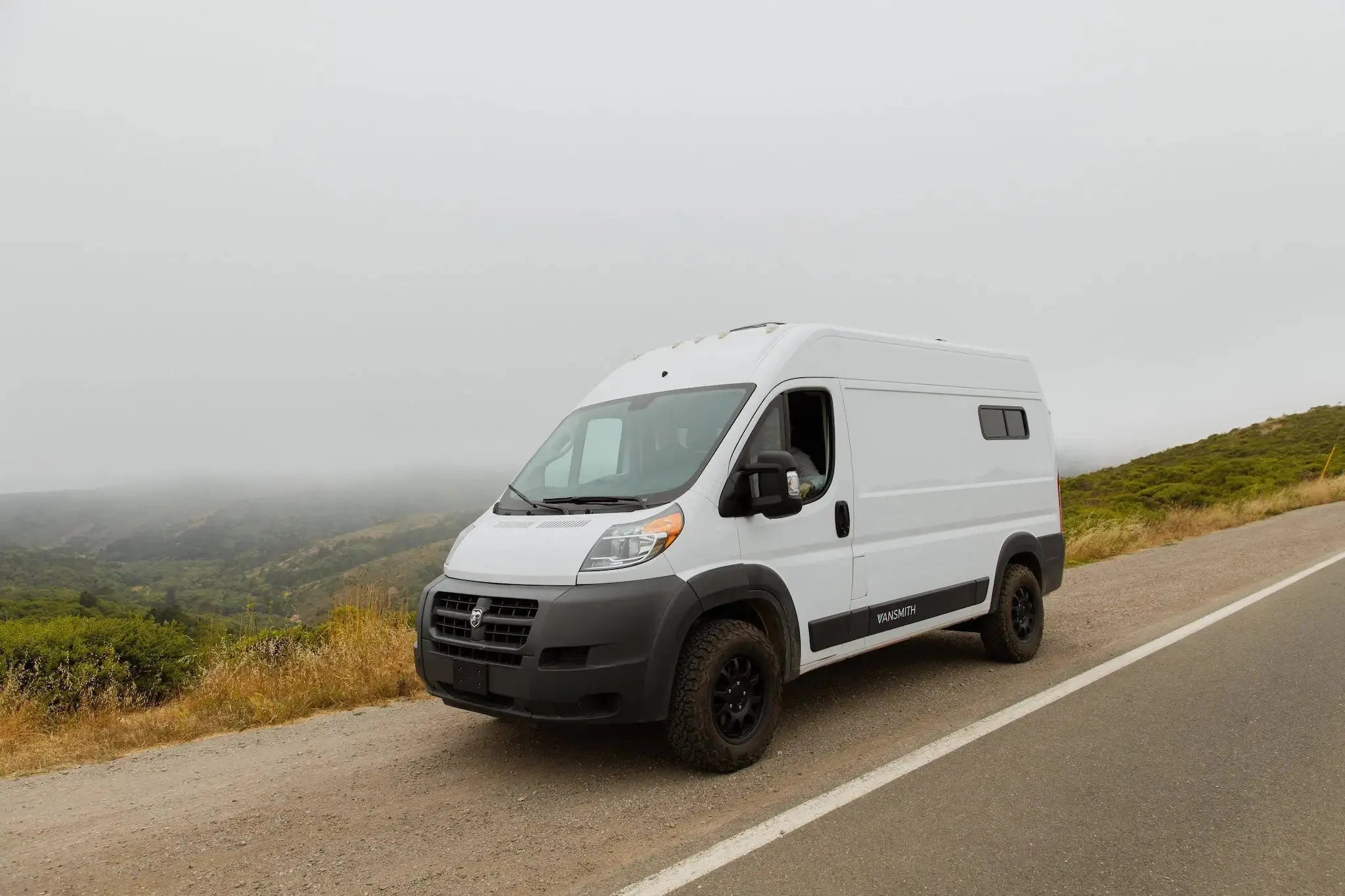 White Ram ProMaster high-roof camper van conversion by The Vansmith, parked roadside at a foggy hillside overlook, black off-road tires and fender flares visible