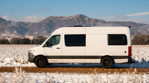 Colorado camper van - This image shows a white Vansmith Sprinter camper van conversion with the Vansmith wordmark on its side, parked on a dirt road beside a snow-dusted field with mountains in the background.