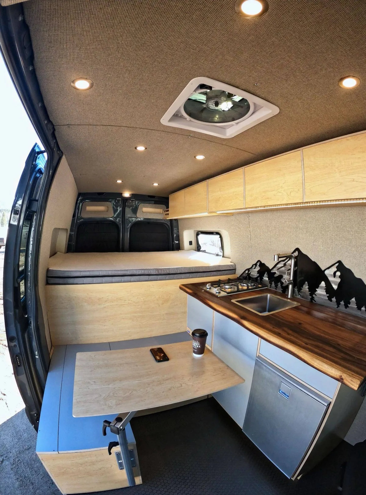 Gina van build interior with gas cooktop, walnut counter, birch cabinets, and elevated bed area