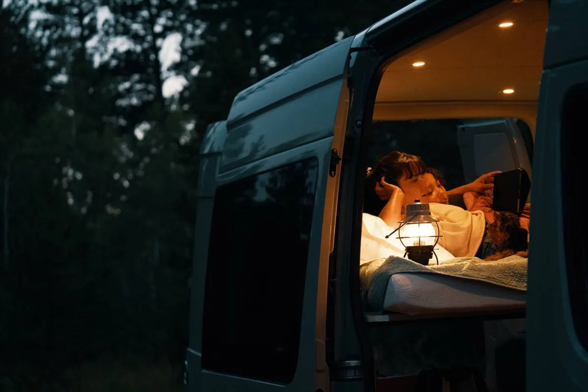 Scout Sprinter van couple reading on bed at night with lantern and sliding door open by The Vansmith